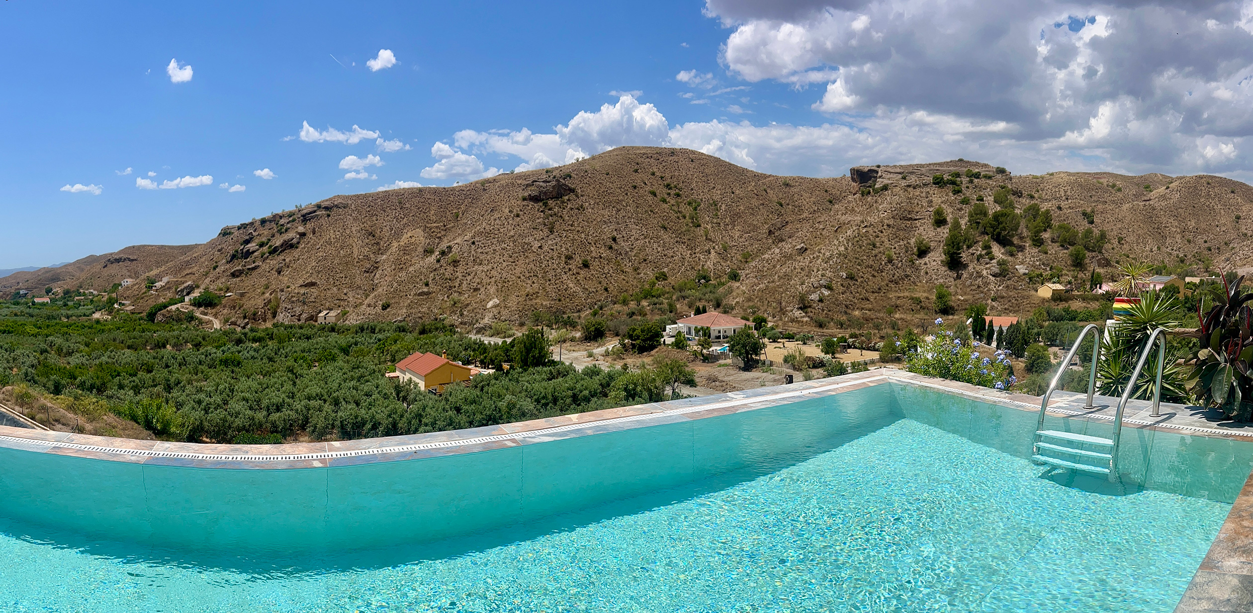 A wide panoramic view of a crystal-clear, turquoise infinity pool at Rambla Retreats, overlooking a green valley and rugged, sunlit desert hills under a partly cloudy blue sky.